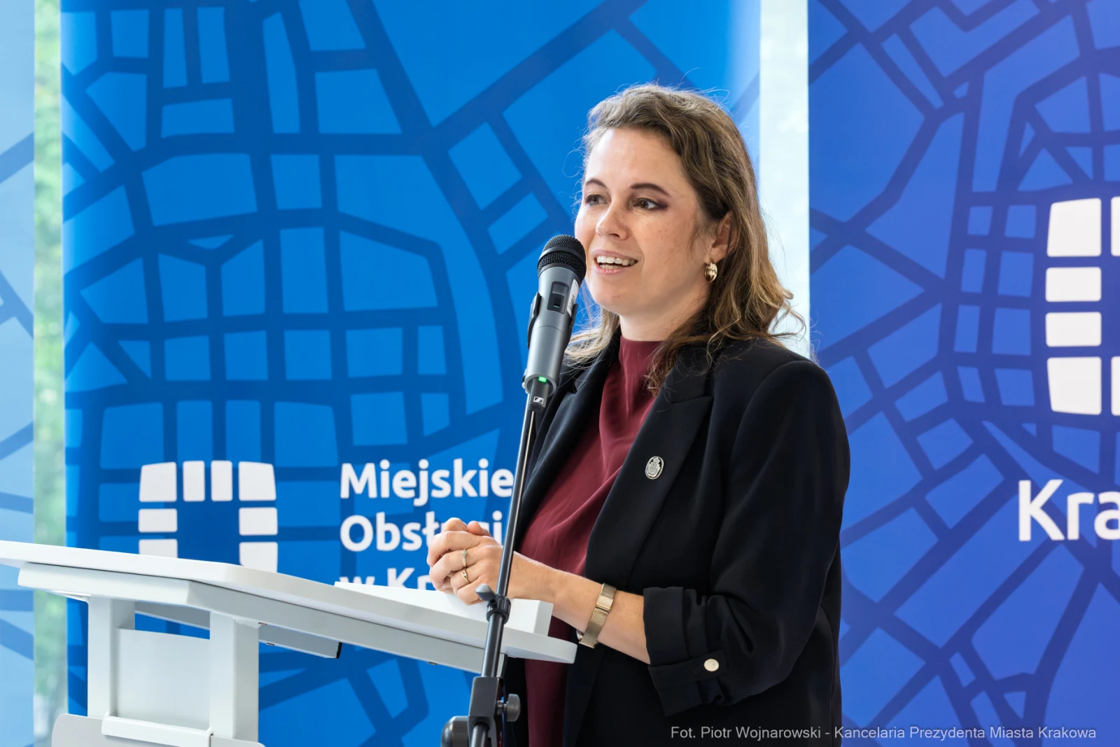 Kobieta przemawia przy mównicy, w tle niebieskie tło z mapą Krakowa.  Autor: Piotr Wojnarowski