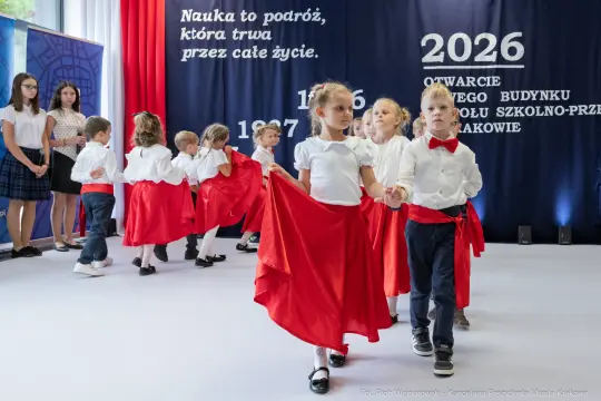 Dzieci w strojach galowych tańczą na scenie podczas uroczystości otwarcia szkoły.