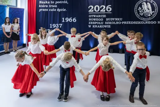 Grupa dzieci w białych koszulach i czerwonych spódnicach tańczy na scenie podczas uroczystości otwarcia.