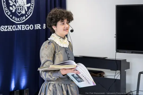 Dziewczyna w szarej sukni z mikrofonem, trzymająca otwartą książkę.