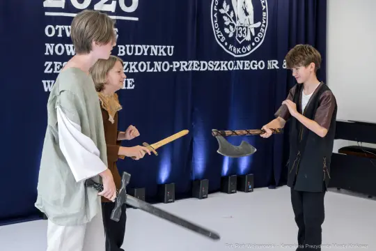 Trzej młodzi ludzie w strojach historycznych, trzymające miecze i topór, w trakcie inscenizacji.