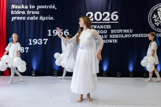 Kobieta w białej sukni tańczy na scenie, obok dzieci z balonami. W tle napisy związane z wydarzeniem.