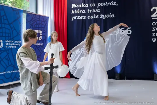 Tancerka w białej sukni wykonuje występ, obok niej stoi chłopiec z mieczem i dziewczynka z balonami.