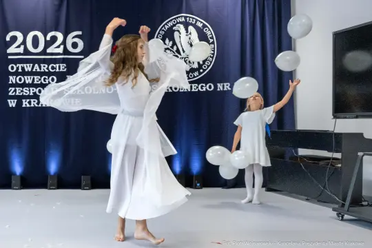 Dwie dziewczynki w białych strojach tańczą z balonami na tle niebieskiego banera.