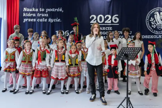 Grupa dzieci w tradycyjnych strojach ludowych występuje na scenie podczas uroczystości.