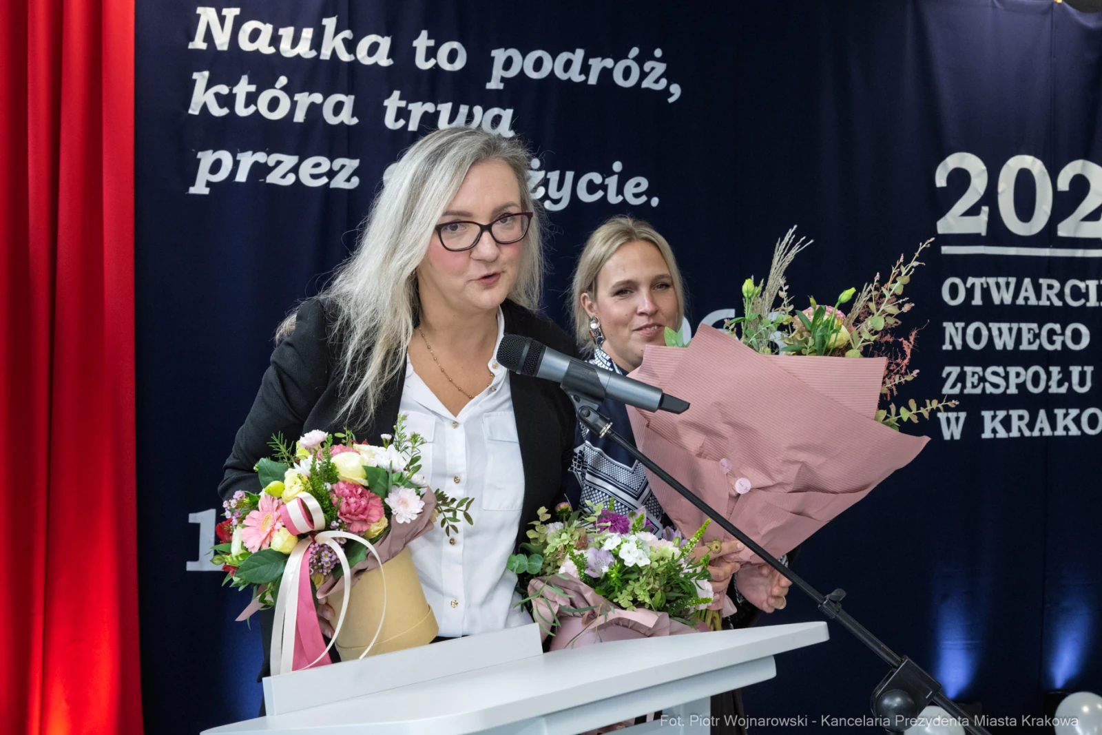 Kobieta przemawia przy mikrofonie, trzymając bukiety kwiatów, obok druga kobieta.  Autor: Piotr Wojnarowski