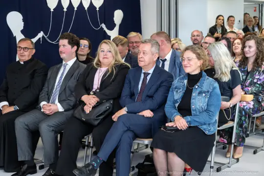 Grupa osób siedzących na wydarzeniu, w tle dekoracja z balonami.
