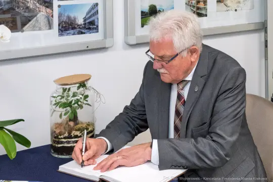 Mężczyzna w garniturze pisze w księdze gości obok rośliny w szklanym słoju.