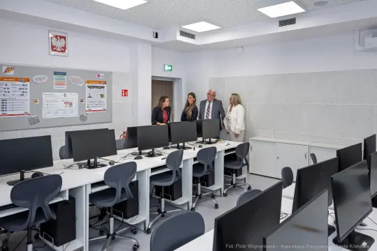 Nowoczesna sala komputerowa z monitorami i grupą osób rozmawiających.