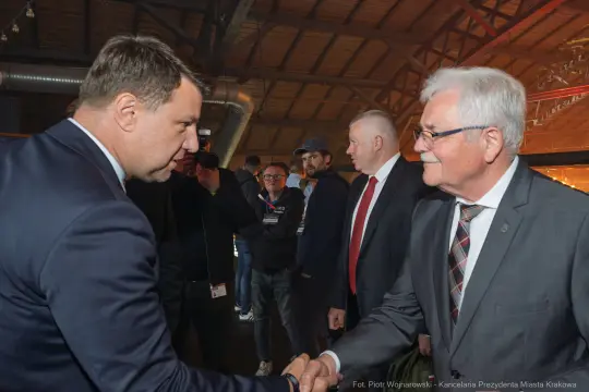 Mężczyźni podają sobie ręce w trakcie spotkania. W tle widoczni inni uczestnicy wydarzenia.