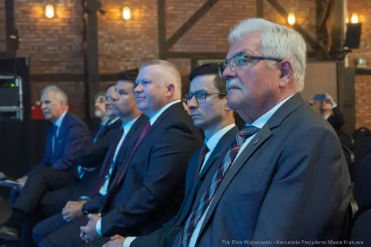 Grupa mężczyzn w garniturach siedzących na wydarzeniu, skupionych na prezentacji.