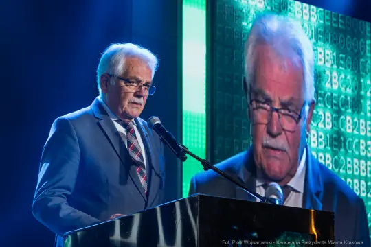 Mężczyzna w okularach przemawia do mikrofonu na tle cyfrowego wyświetlacza.