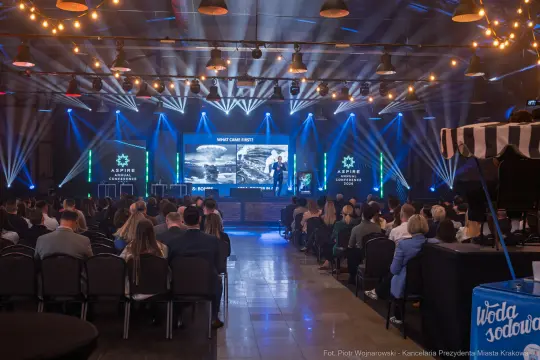 Konferencja z prezentacją na dużym ekranie, oświetlenie sceniczne, publiczność siedząca na krzesłach.