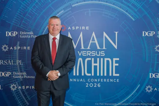 Mężczyzna w garniturze stoi na tle logo konferencji Man versus Machine 2026.