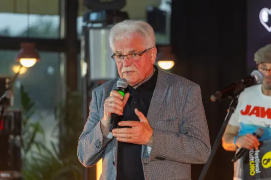 Mężczyzna w okularach, w garniturze, przemawia do mikrofonu na scenie.