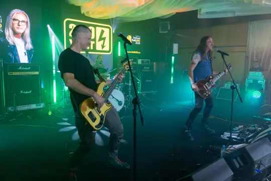 Zespół rockowy grający na scenie, z efektami świetlnymi i sprzętem muzycznym w tle.