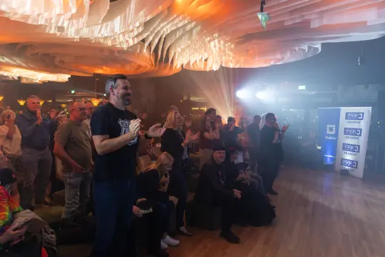 Ludzie na wydarzeniu stoją i klaskają, w tle oświetlenie i scena.