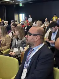 Grupa ludzi siedzących na konferencji, niektórzy z telefonami i notatkami.