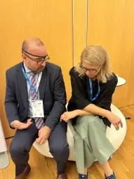 Dwoje ludzi w formalnych ubraniach rozmawia ze sobą, siedząc na okrągłej kanapie.