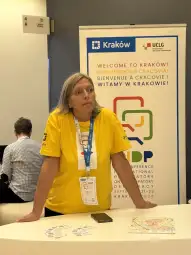 Kobieta w żółtej koszulce stoi przy stoisku konferencyjnym w Krakowie.