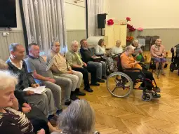 Grupa seniorów siedzących w sali, część z nich na wózkach inwalidzkich.