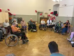 Spotkanie osób starszych w sali, niektórzy na wózkach inwalidzkich, grają muzykę i śpiewają.