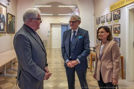 Trzech ludzi rozmawia w korytarzu z portretami na ścianach.