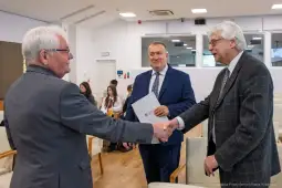 Trzech mężczyzn w formalnych strojach, uściski dłoni i rozmowy w sali konferencyjnej.