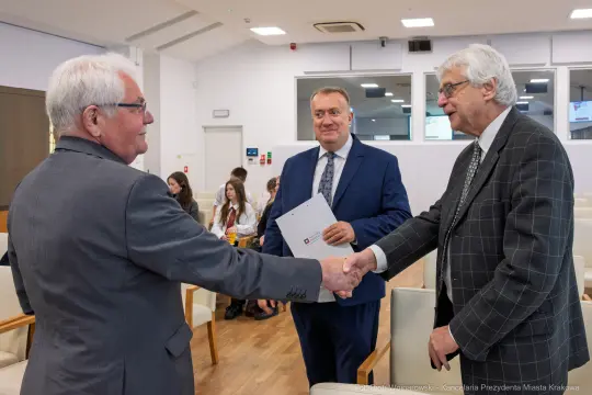 Trzech mężczyzn w formalnych strojach, uściski dłoni i rozmowy w sali konferencyjnej.
