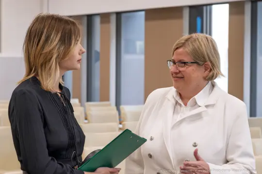 Dwie kobiety rozmawiają w sali konferencyjnej. Jedna trzyma teczkę.