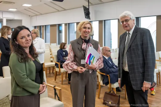 Trzy osoby w formalnych strojach rozmawiają w nowoczesnej sali konferencyjnej.