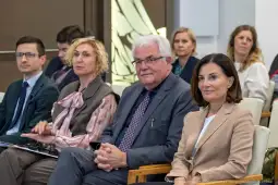 Grupa osób siedzących na spotkaniu, w tle widoczni uczestnicy w formalnych strojach.