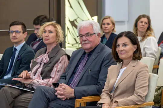 Grupa osób siedzących na spotkaniu, w tle widoczni uczestnicy w formalnych strojach.