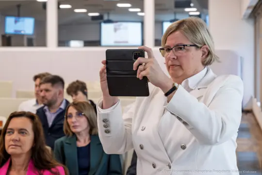Kobieta robiąca zdjęcie telefonem, w tle grupa osób siedzących na sali.