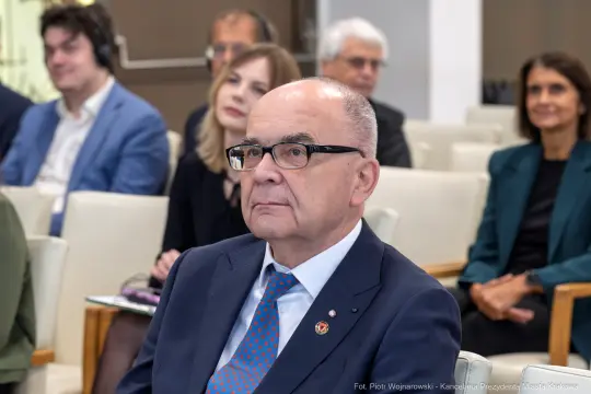 Mężczyzna w okularach, w garniturze, słucha wystąpienia na konferencji. W tle inni uczestnicy.
