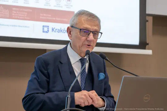 Mężczyzna w okularach przemawia do mikrofonu, obok laptop i ekran z prezentacją.
