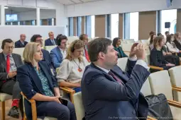 Grupa ludzi siedzących w sali konferencyjnej, niektórzy z telefonami w rękach.
