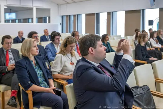 Grupa ludzi siedzących w sali konferencyjnej, niektórzy z telefonami w rękach.