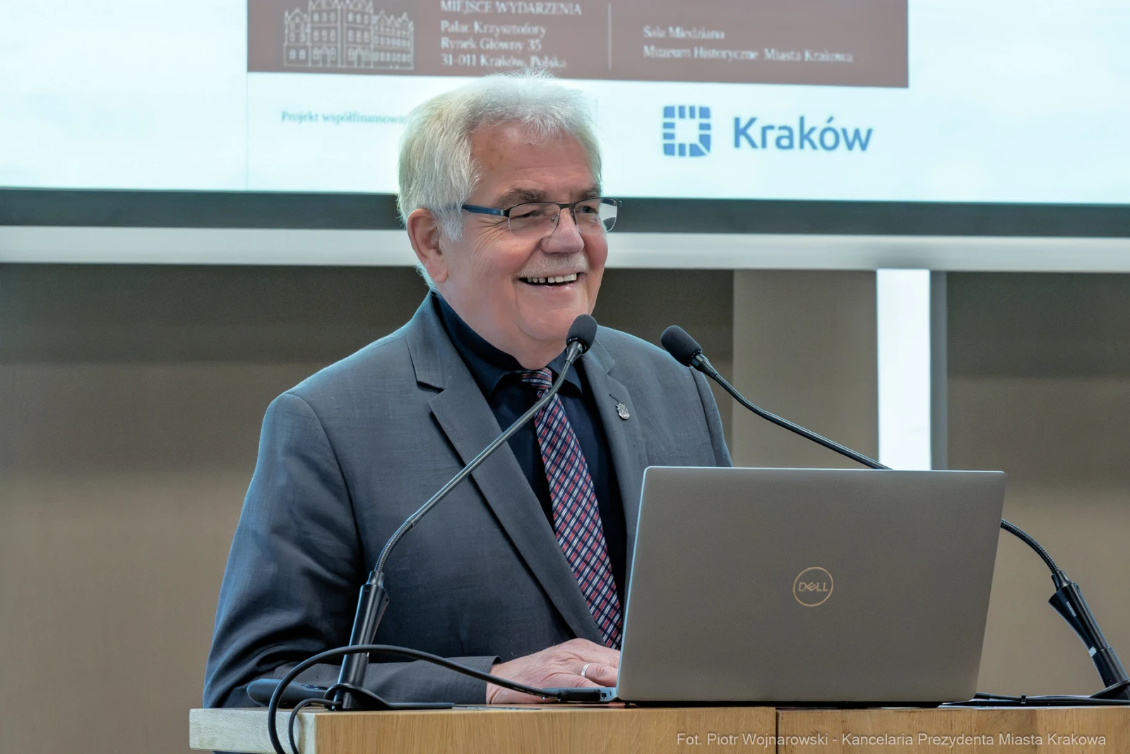 Mężczyzna w garniturze uśmiecha się, stojąc przy mównicy z laptopem.  Autor: Piotr Wojnarowski