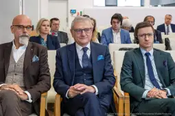 Trzech mężczyzn w garniturach siedzi w sali konferencyjnej, w tle inni uczestnicy spotkania.
