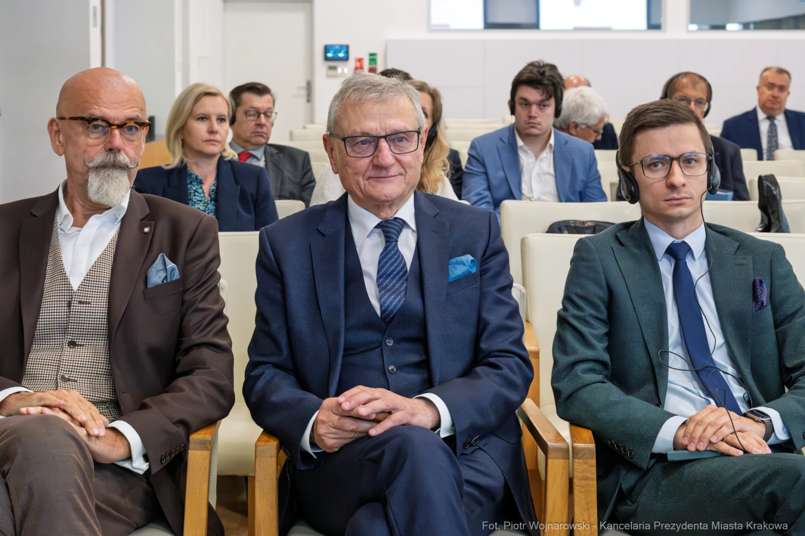 Trzech mężczyzn w garniturach siedzi w sali konferencyjnej, w tle inni uczestnicy spotkania.  Autor: Piotr Wojnarowski