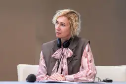 Kobieta z kręconymi włosami w bluzce z kokardą, siedząca przy stole z mikrofonem.