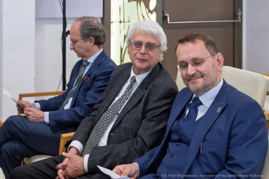 Trzech mężczyzn w garniturach siedzi obok siebie, uśmiechając się i trzymając dokumenty.