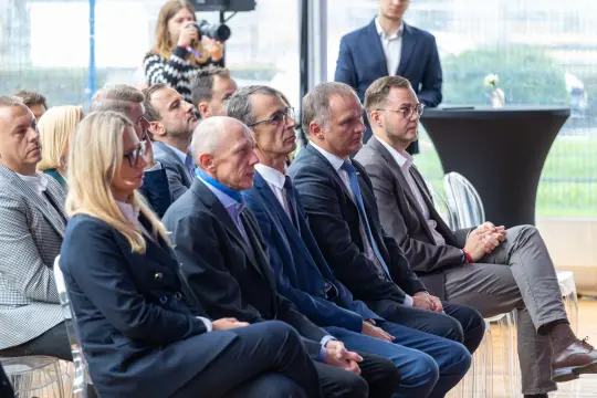 Grupa mężczyzn i kobiet siedzących w sali konferencyjnej, skupionych na wydarzeniu.