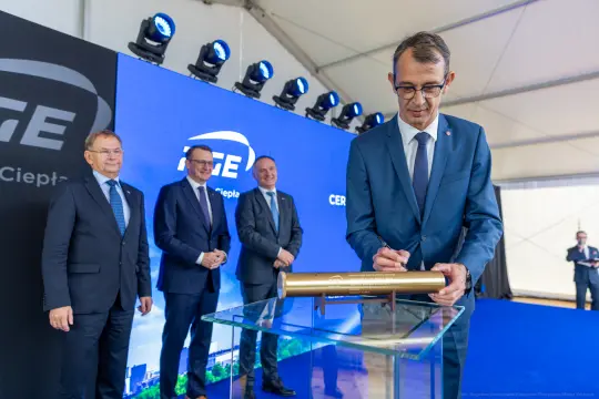Ceremonia podpisania dokumentu przez mężczyznę w garniturze, obok trzech innych mężczyzn.
