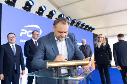 Mężczyzna w garniturze podpisuje złotą tablicę podczas wydarzenia. W tle logo firmy i zgromadzeni goście.