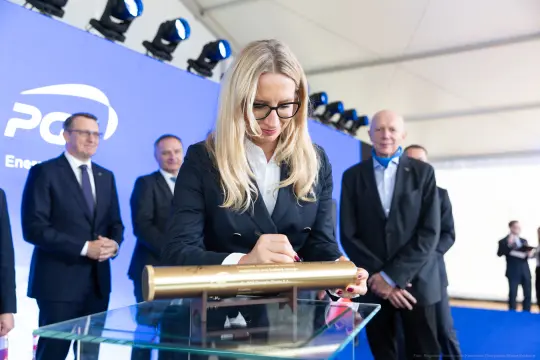 Kobieta podpisuje złotą tubę w otoczeniu mężczyzn na tle niebieskiego banera.