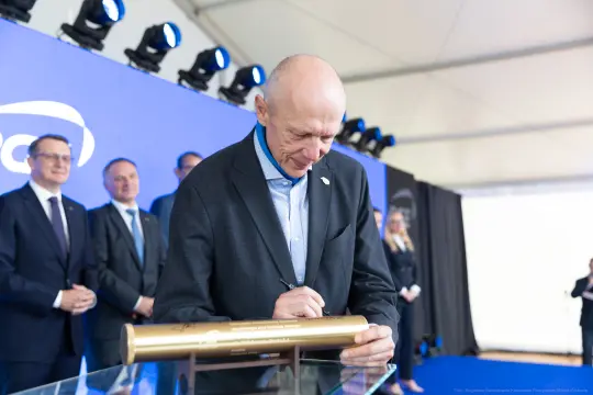 Mężczyzna podpisuje złotą tubę na tle niebieskiej ścianki z oświetleniem.