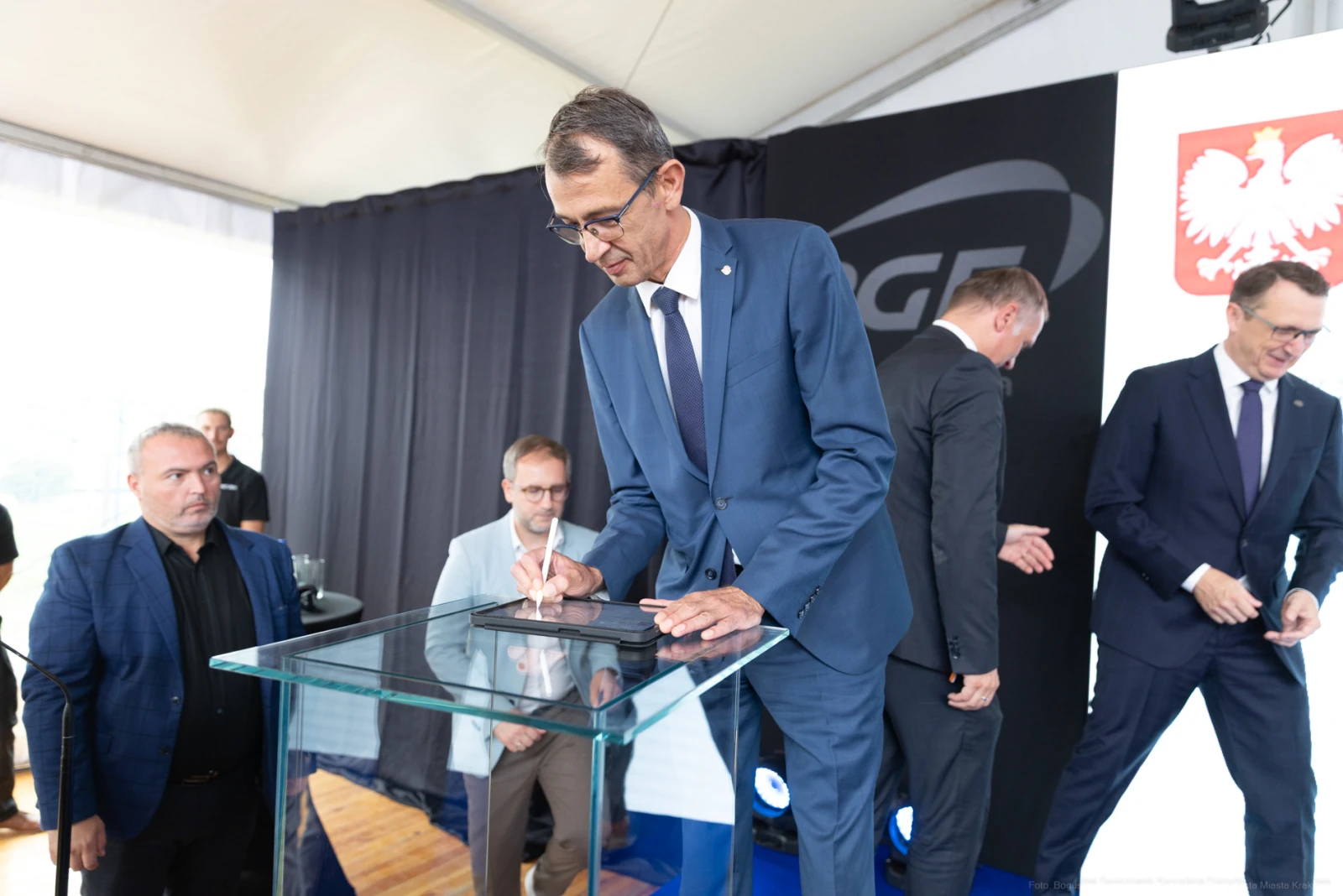 Mężczyzna podpisujący dokument na szklanym stole, w tle inni uczestnicy wydarzenia.  Autor: Bogusław Świerzowski