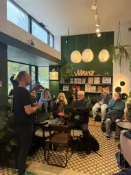 Spotkanie w kawiarni z grupą ludzi słuchających prelegenta. Rośliny w tle.